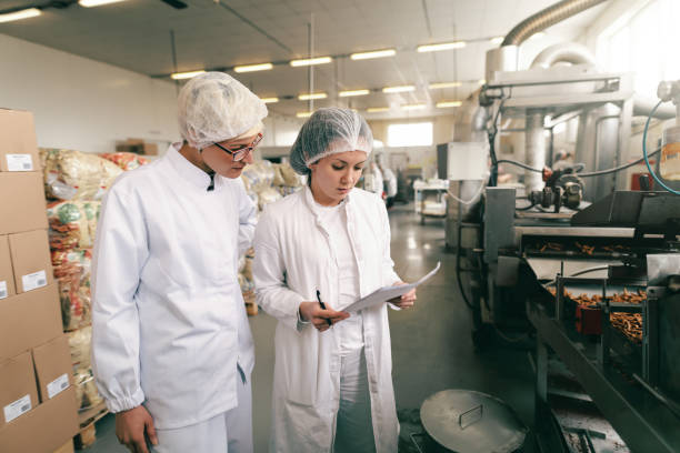 Two quality professionals in white sterile uniforms checking quality of food while standing in food manufacturing factory