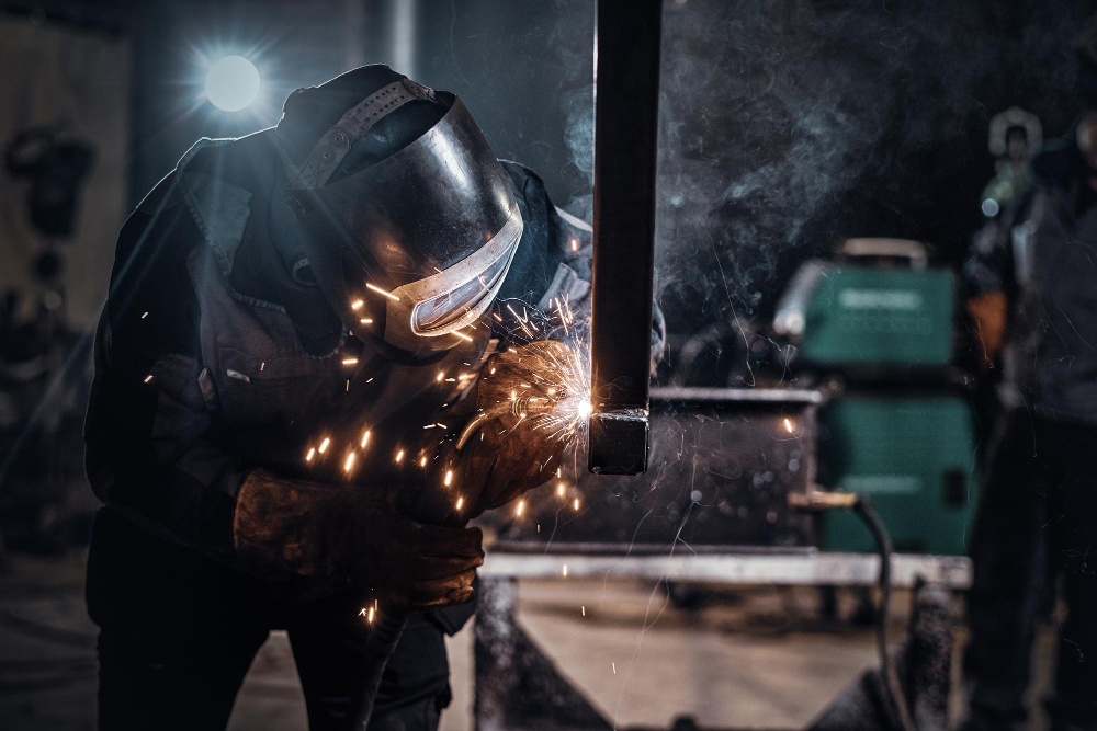 A welder in protective gear expertly creates sparks while working on metal in a dimly lit workshop, surrounded by machinery