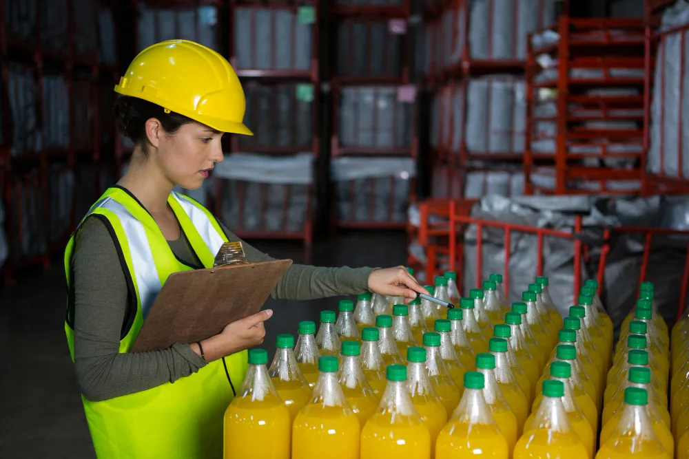 Beverage production quality control inspection of bottled juice by a female worker in a warehouse setting Female worker conducting beverage production quality control by inspecting bottled juice in a warehouse while holding a clipboard and wearing safety gear