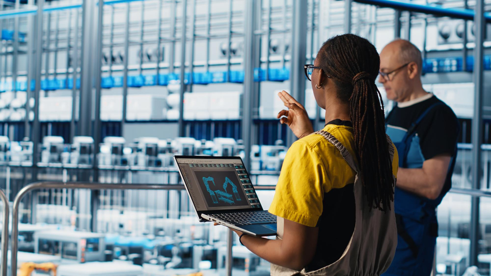 Engineer using laptop with 3D model in a smart factory environment, discussing automation processes with technician in high-tech manufacturing facility